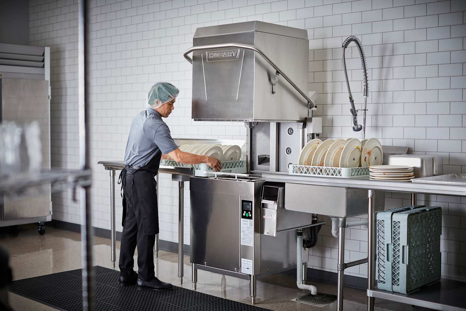 A kitchen worker operates a Hobart commercial door-type dishwasher at a professional dishwashing station