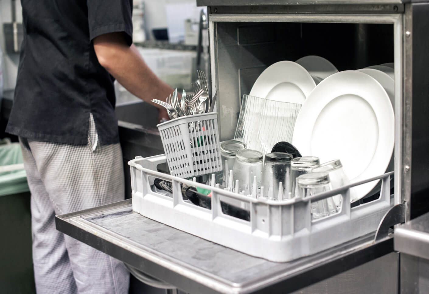 Clean dishes and cutlery are loaded into a commercial dishwasher in a busy kitchen