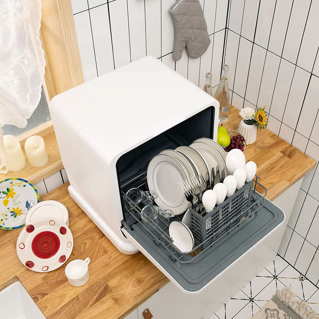 A compact white benchtop dishwasher with its rack pulled out, showing loaded dishes and cutlery, set on a wooden kitchen counter
