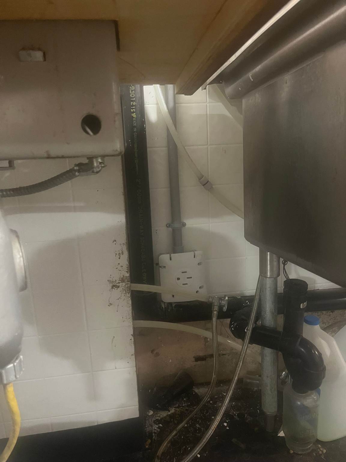 Plumbing and electrical setup under a commercial dishwasher showing water lines, drainage, and power outlet connections